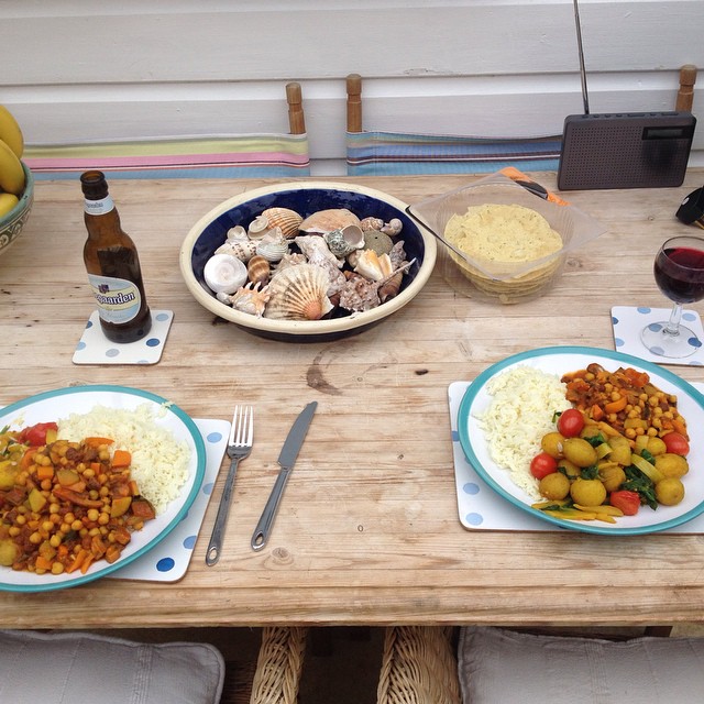 Wooden table set with two plates of rice and vegetable stew, a beer bottle, wine glass, bowl of shells, and radio.
