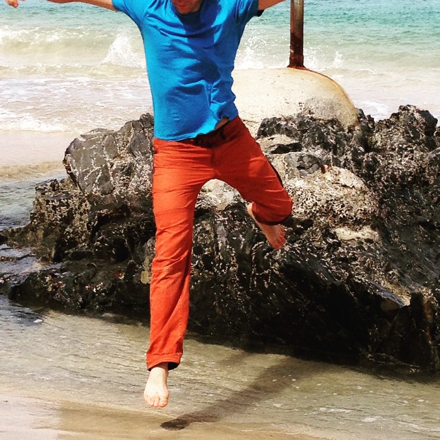 Barefoot person in blue shirt and orange pants jumping mid-air near rocks and ocean waves on a beach.