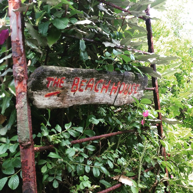 Wooden sign reading "The Beach House" painted in red, mounted on a rusted metal gate surrounded by green foliage.