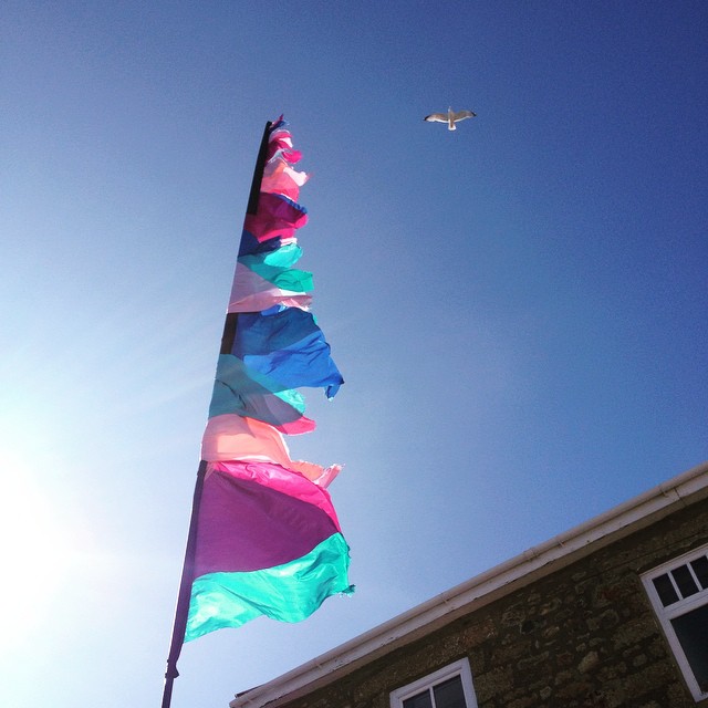 Colorful fabric streamers on a pole against blue sky, sun glare, seagull flying, stone building with white window trim below.