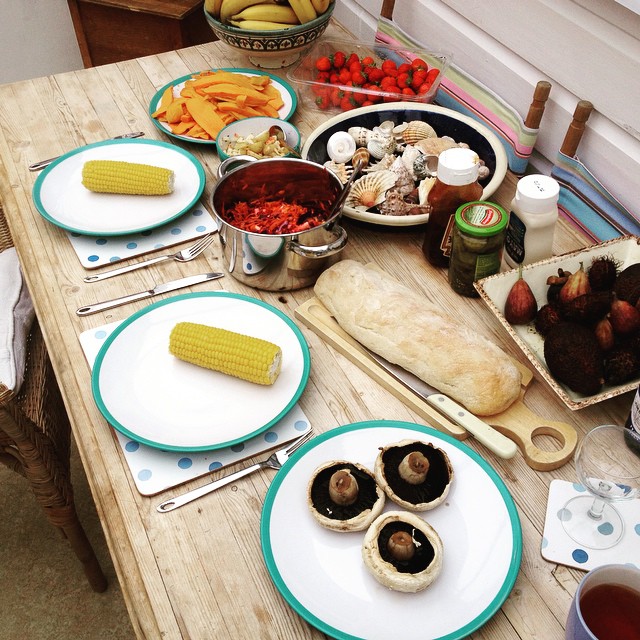 Wooden table set with corn on plates, mushrooms, bread, seashells, fruit, and condiments.