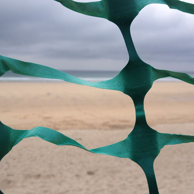 Green plastic mesh fence in foreground with sandy beach and overcast sky visible through gaps.