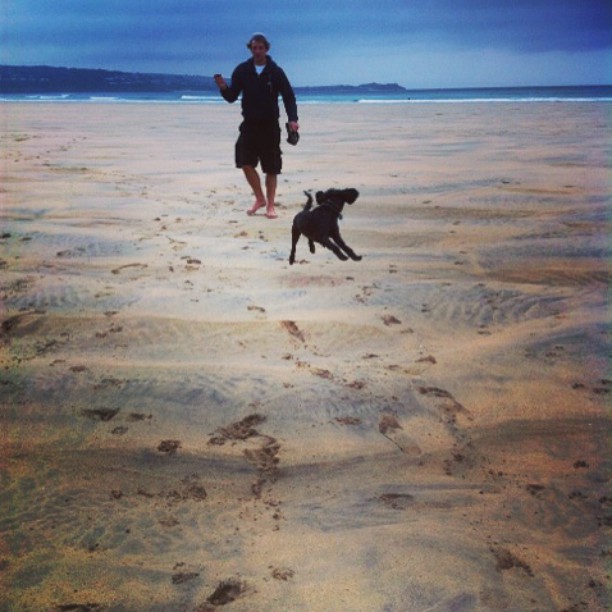Man and dog running on a sandy beach, footprints trailing behind, ocean and hazy coastline in the background.