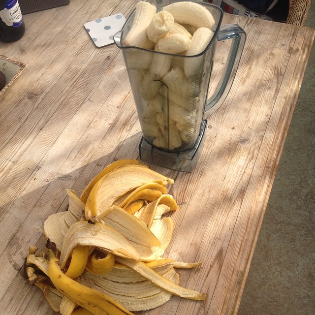 Blender pitcher filled with peeled bananas next to a pile of banana peels on a wooden table.