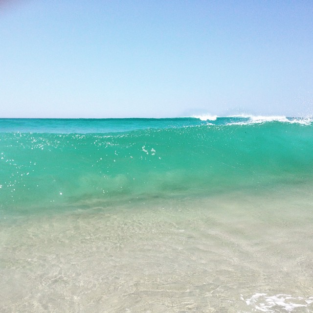 Clear turquoise ocean wave curling near shore, shallow sandy water in foreground, pale blue sky above.