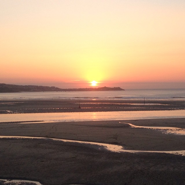 Sun setting low over the sea, orange sky, distant coastline silhouette, wet sand and tidal pools in foreground.