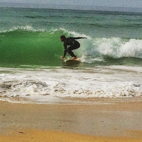Surfer in wetsuit riding a small breaking wave near shore, with sandy beach in the foreground.