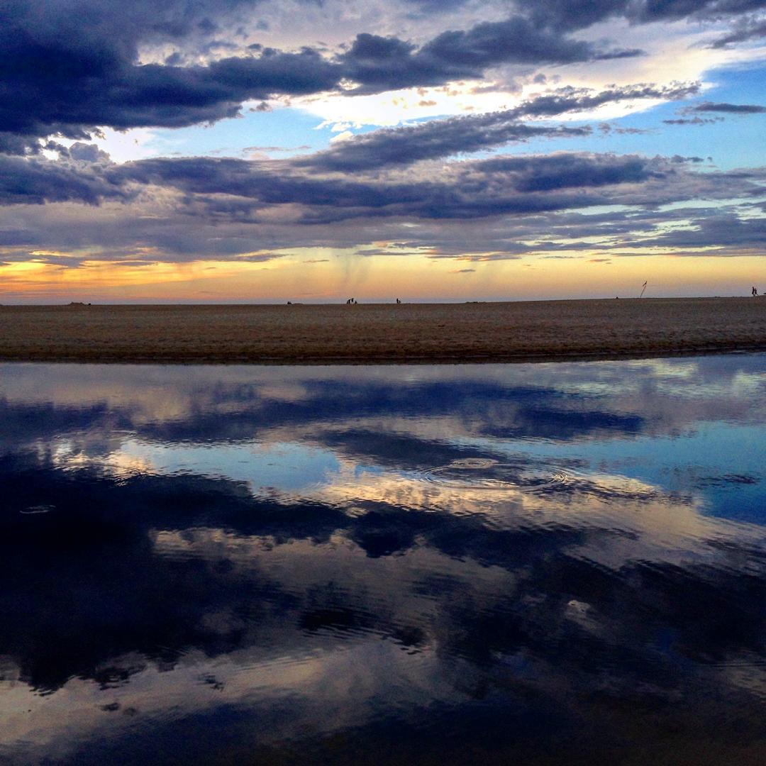 Beach at dusk with dramatic clouds reflected in still water on wet sand, distant figures on the shore.