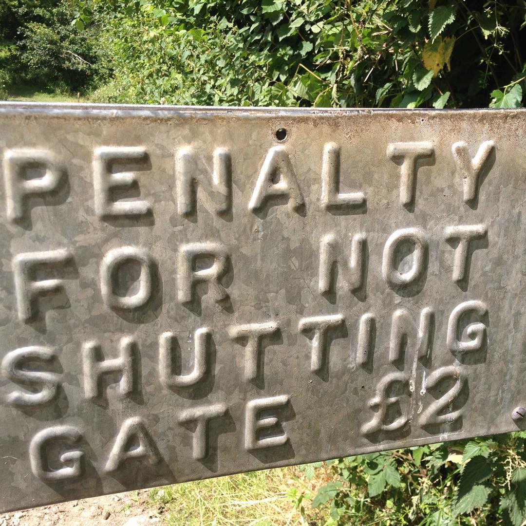 Weathered metal sign with embossed letters reading "PENALTY FOR NOT SHUTTING GATE £2," surrounded by green foliage.