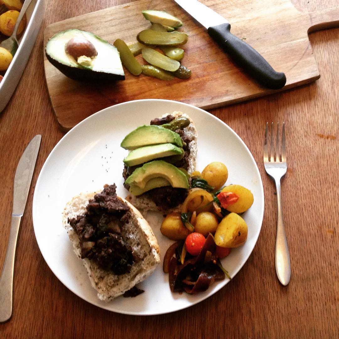 Overhead view of open sandwiches with avocado slices and minced filling on bread, potatoes and tomatoes, beside a cutting board with sliced avocado and pickles.