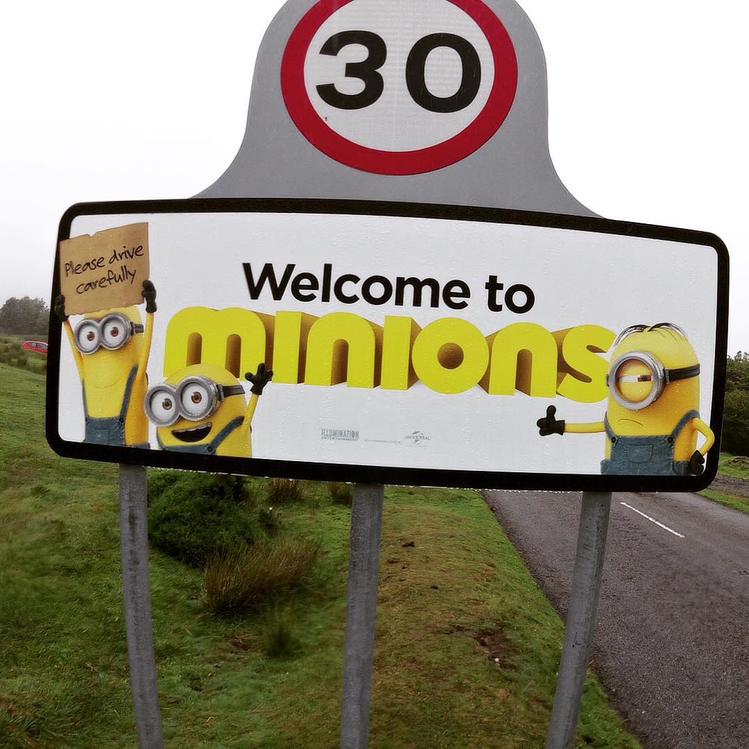Road sign reading "Welcome to Minions" with cartoon Minion characters, below a 30 mph speed limit sign, grassy roadside setting.
