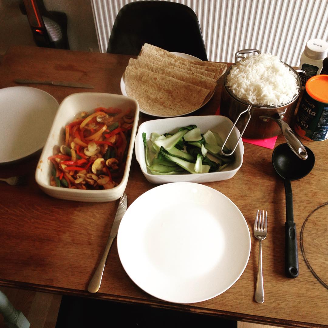 Table set with empty plate, fork, and knife, surrounded by dishes of rice, tortillas, sautéed peppers and mushrooms, and bok choy.