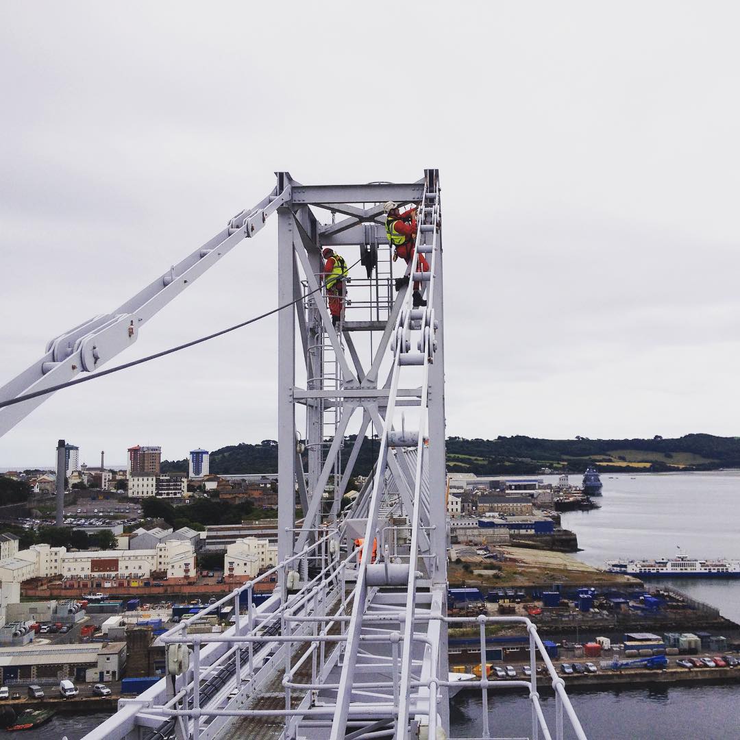 Workers in harnesses on a suspension bridge tower, with a coastal town and harbor visible below.