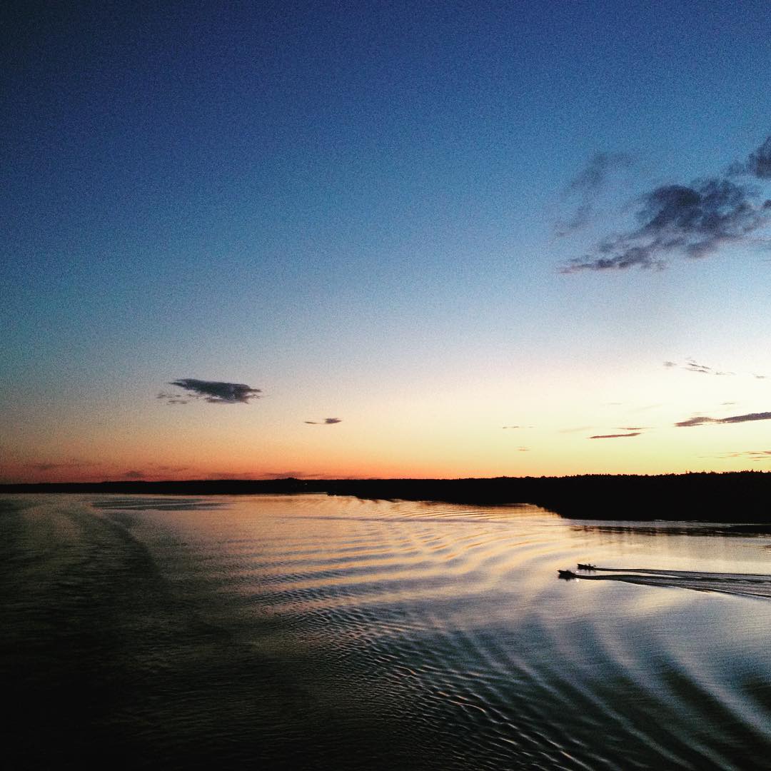 Small boat leaving a wake on calm water at dusk, with a dark tree-lined shoreline and gradient sky from orange horizon to blue above.