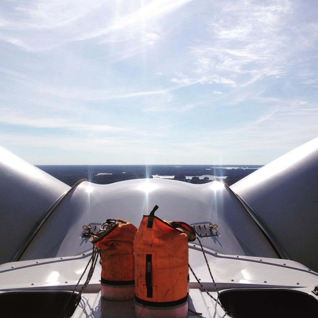 Two orange gear bags atop a wind turbine nacelle, with turbine blades framing a wide aerial view of forested land and lakes below.
