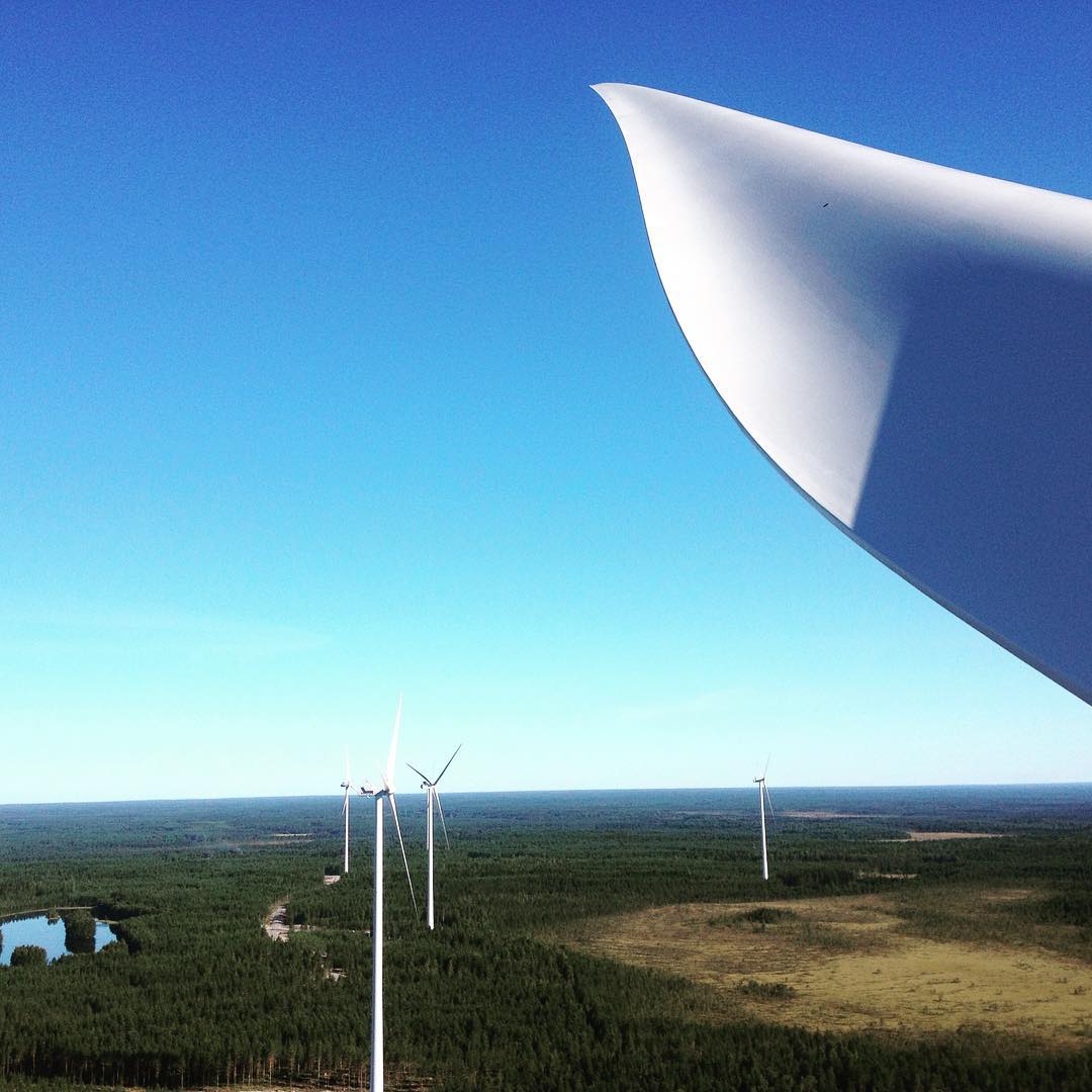 Close-up of a wind turbine blade against a blue sky, overlooking a forested landscape with several distant wind turbines and a small lake.