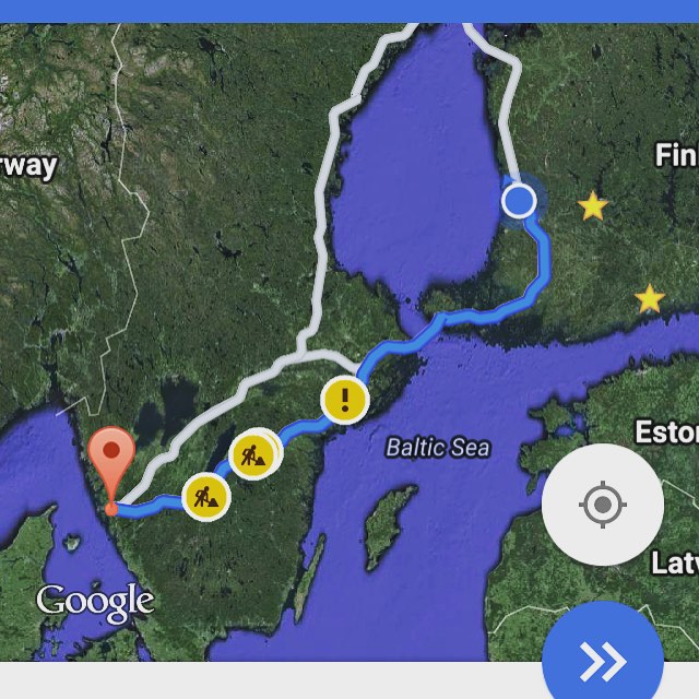 Satellite map view showing a blue navigation route with two warning icons and a location pin, spanning Scandinavia and the Baltic Sea.