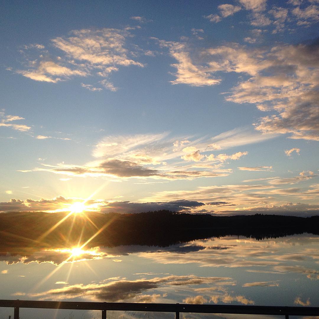 Sunset over a still lake with sun star on horizon, silhouetted hills, reflected clouds, and a railing in foreground.