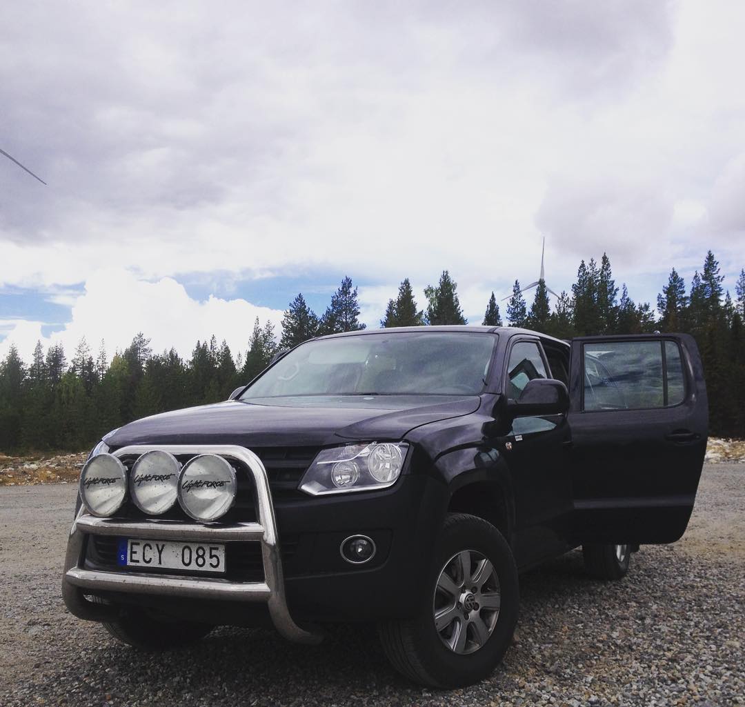 Black pickup truck with bull bar and auxiliary lights parked on gravel, doors open, forest and wind turbine in background.
