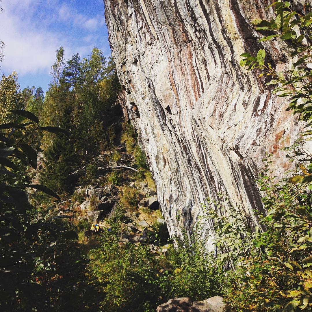 Steep striated rock cliff face beside a forested hillside with scattered boulders and green undergrowth under a blue sky with clouds.
