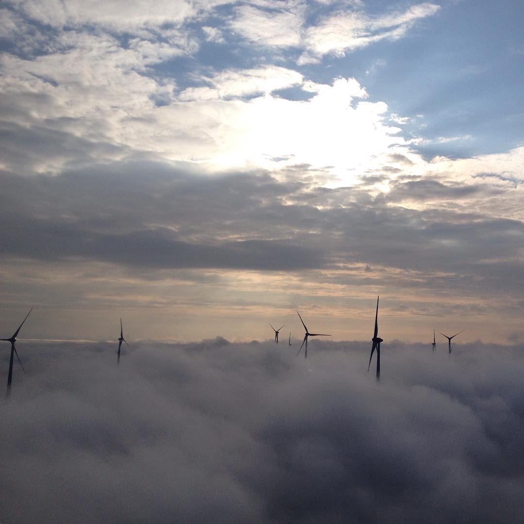 Wind turbines rising above a thick layer of clouds, silhouetted against a sky with sunlight breaking through clouds above.
