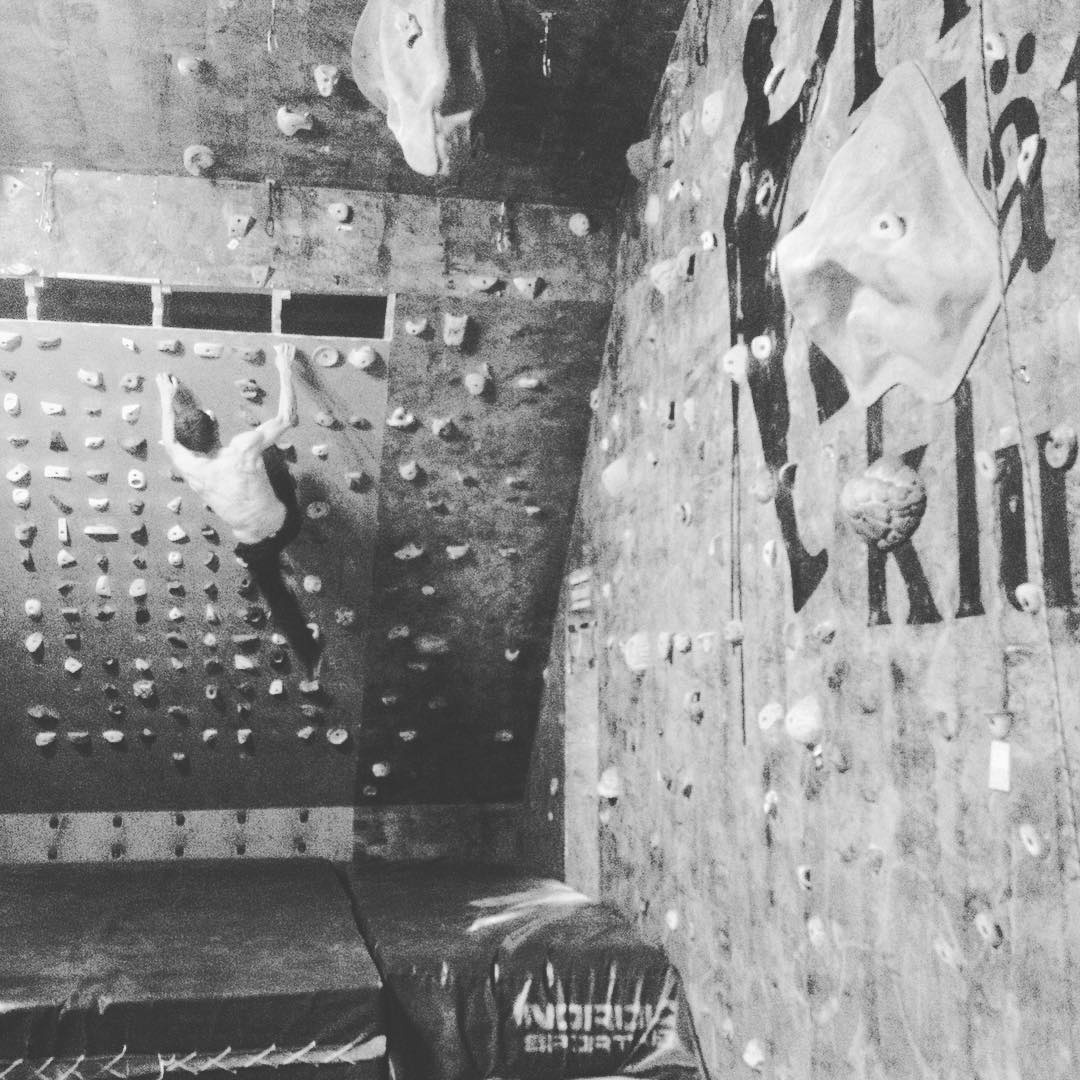 Black and white photo of a person climbing an indoor bouldering wall covered with colorful climbing holds, crash pads below.