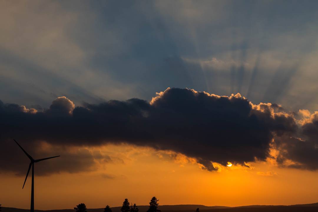 Silhouette of a wind turbine and trees against a sunset, with sun rays fanning out behind dark clouds.