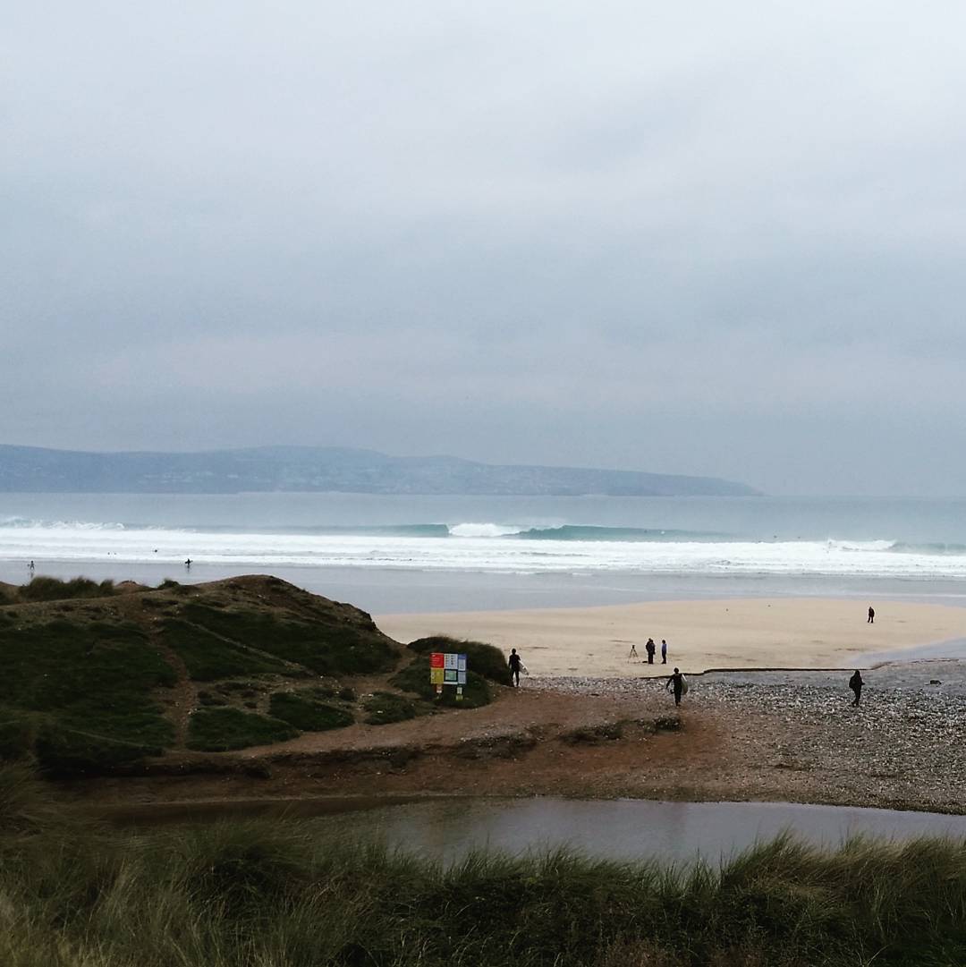 Wide sandy beach with breaking waves, distant headland, overcast sky, and a few people walking near dunes and a small stream.