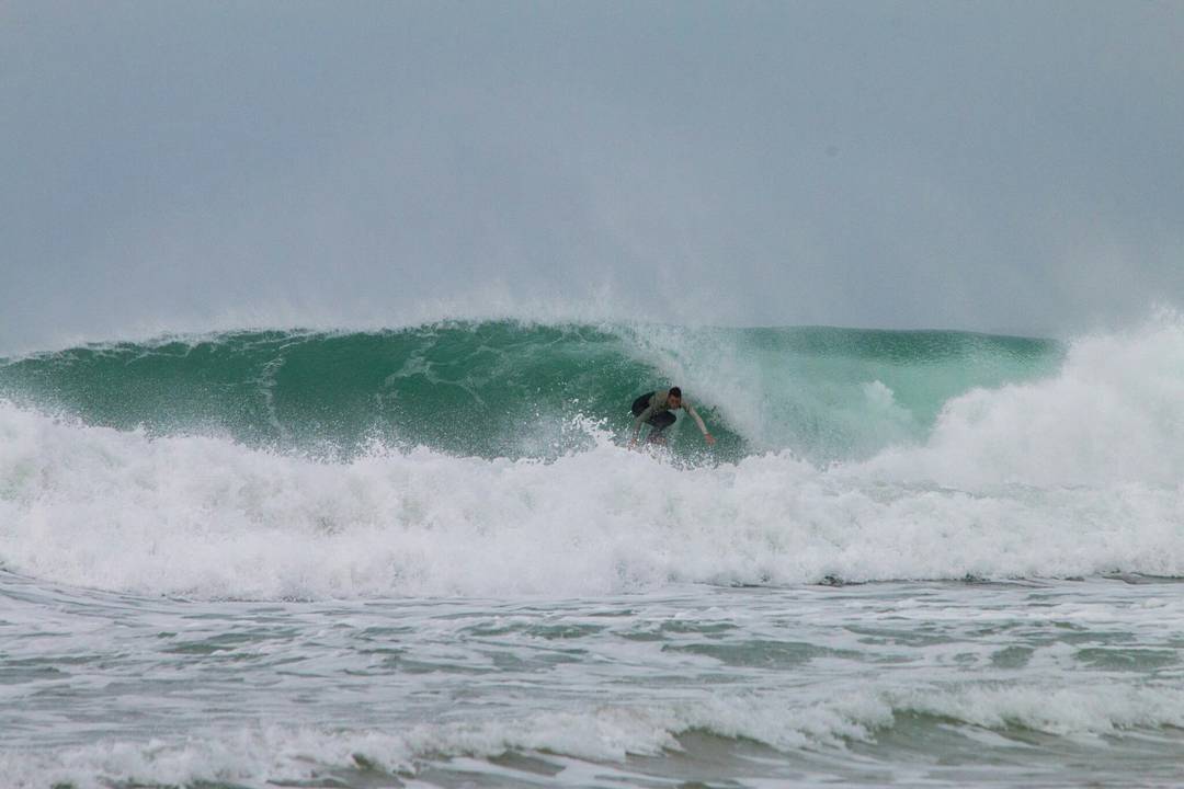 Surfer crouched inside a curling green wave, surrounded by white spray, under an overcast sky.