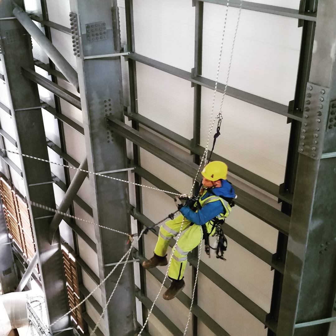 Rope access technician in yellow helmet and hi-vis suit suspended on ropes inside a steel-framed industrial structure.