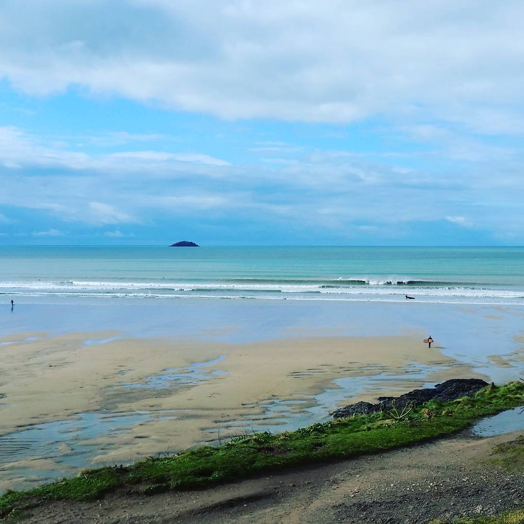 Wide sandy beach at low tide with distant surfers, gentle waves, a small island on the horizon, and a grassy foreground under a cloudy sky.