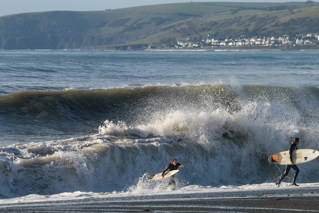 Two surfers walking on a beach with surfboards as a large wave breaks behind them, hills and coastal town in background.