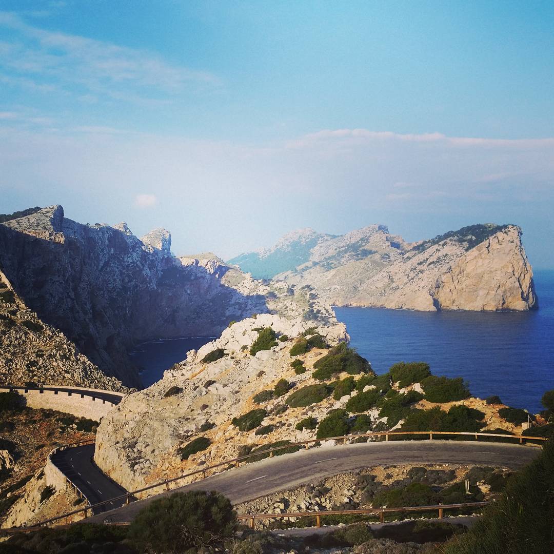 Winding coastal road curving along rocky cliffs above a blue bay, with rugged mountain headlands in the background under a clear sky.