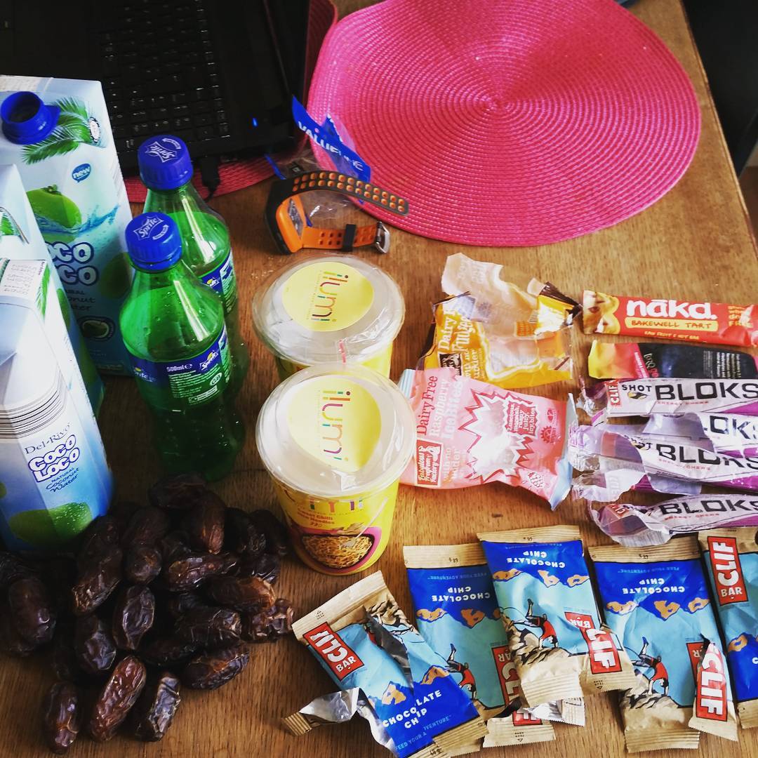 Table with coconut water carton, soda bottles, dates, instant noodle cups, energy bars (Clif, Nakd), gel packs, a pink hat, and a sports watch.