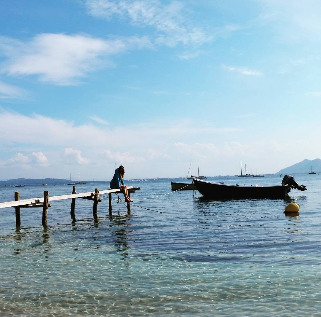 Person sitting on a wooden pier over clear shallow water, boats anchored nearby, distant hills and cloudy blue sky.