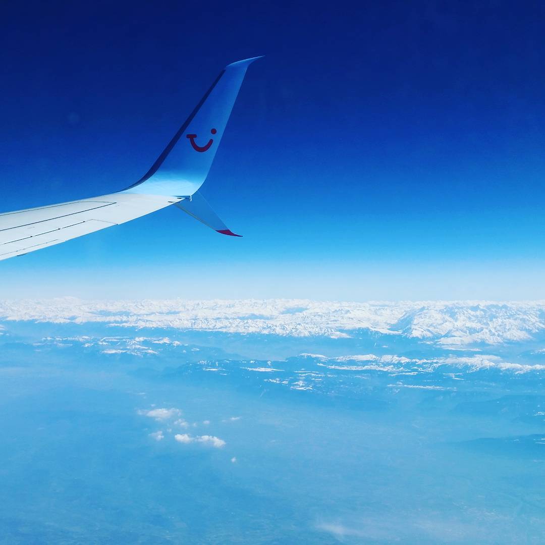 Airplane wing with TUI logo against deep blue sky, snow-capped mountain range visible below through thin clouds.