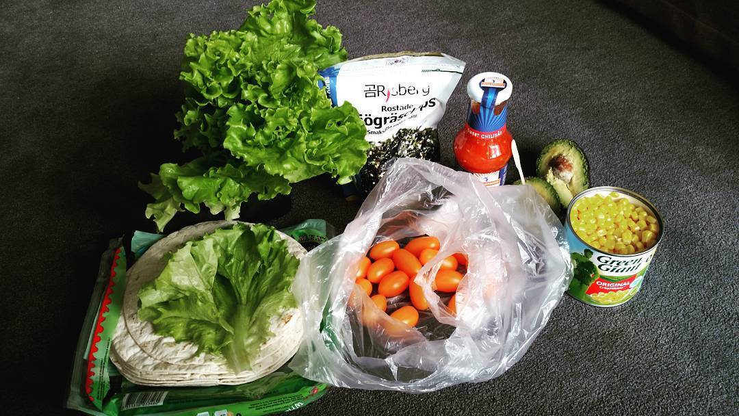 Groceries on carpet: lettuce, tortillas, cherry tomatoes in a bag, canned corn, seaweed snacks, chili sauce, and an avocado.