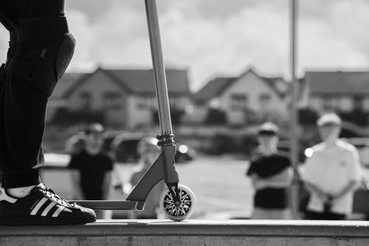 Black-and-white photo of a stunt scooter's front wheel on a ramp edge, with a person's foot in sneakers nearby and onlookers blurred in background.