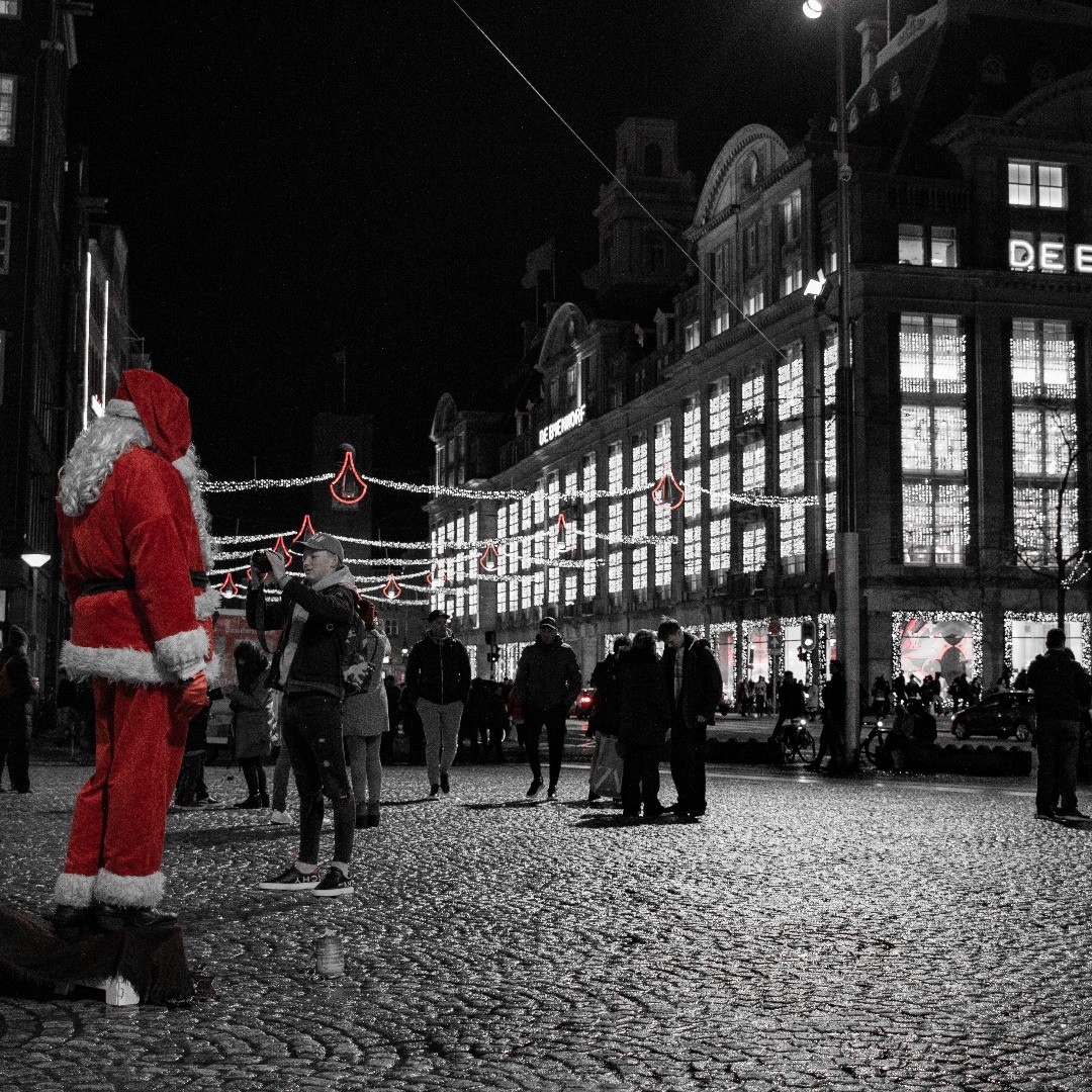 Black-and-white night photo of a city square with a person in a red Santa costume standing on a box while a crowd watches, illuminated storefront behind.