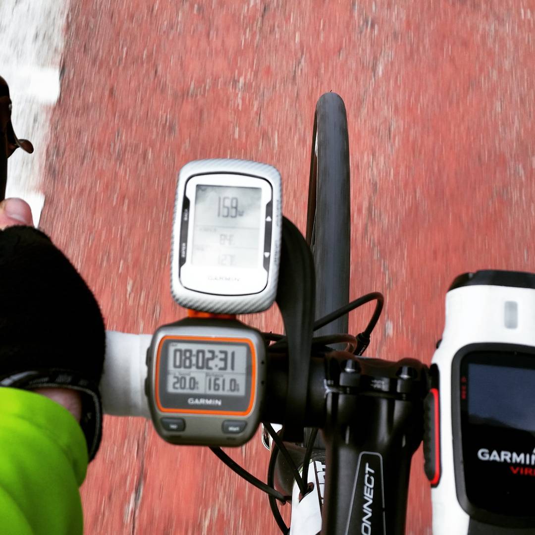 Cyclist's handlebar view with two Garmin GPS devices showing power, time, and distance data, over a red road surface.