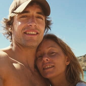 Close-up selfie of a man in a cap and a woman with brown hair smiling, outdoors near water under a blue sky.
