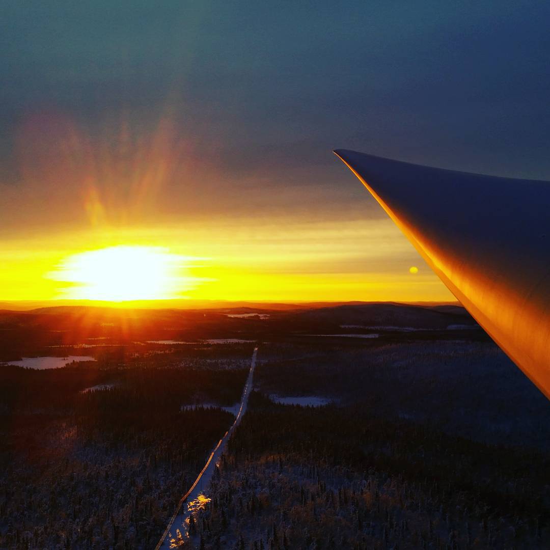 Aerial view from an aircraft wing at sunset, with a bright orange sun on the horizon over a snowy forested landscape and a straight road below.