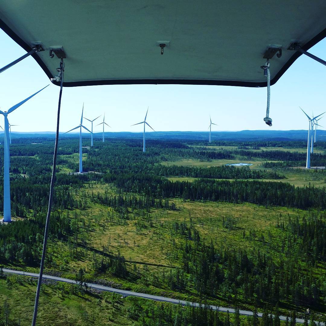 Aerial view from beneath a helicopter or gondola roof overlooking a wind farm amid forested land with a road below.
