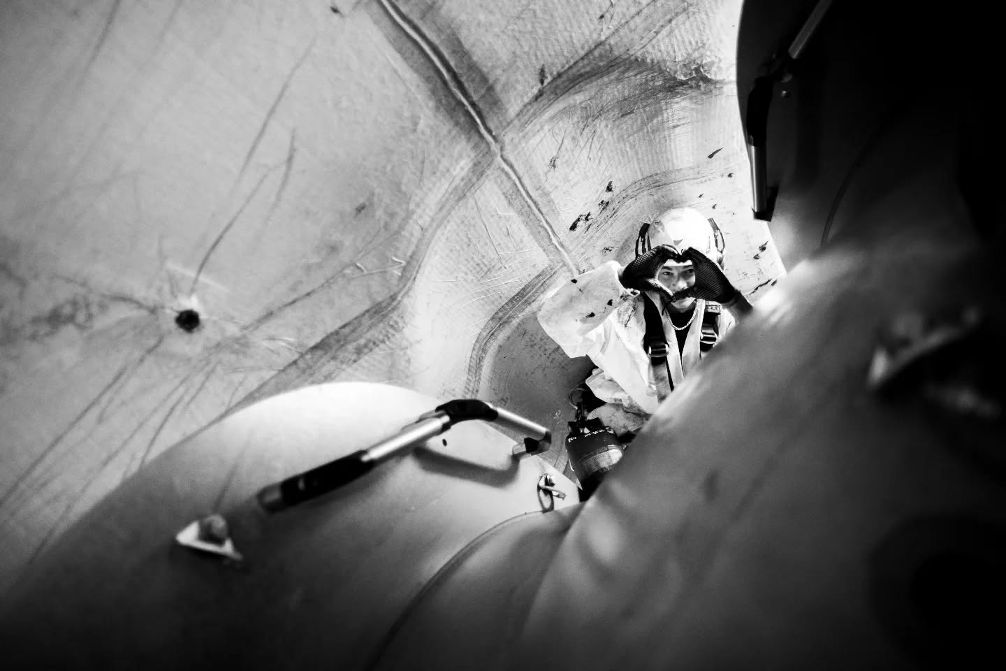 Black and white photo of a worker in protective suit and helmet inside a wind turbine blade, making a heart shape with hands.