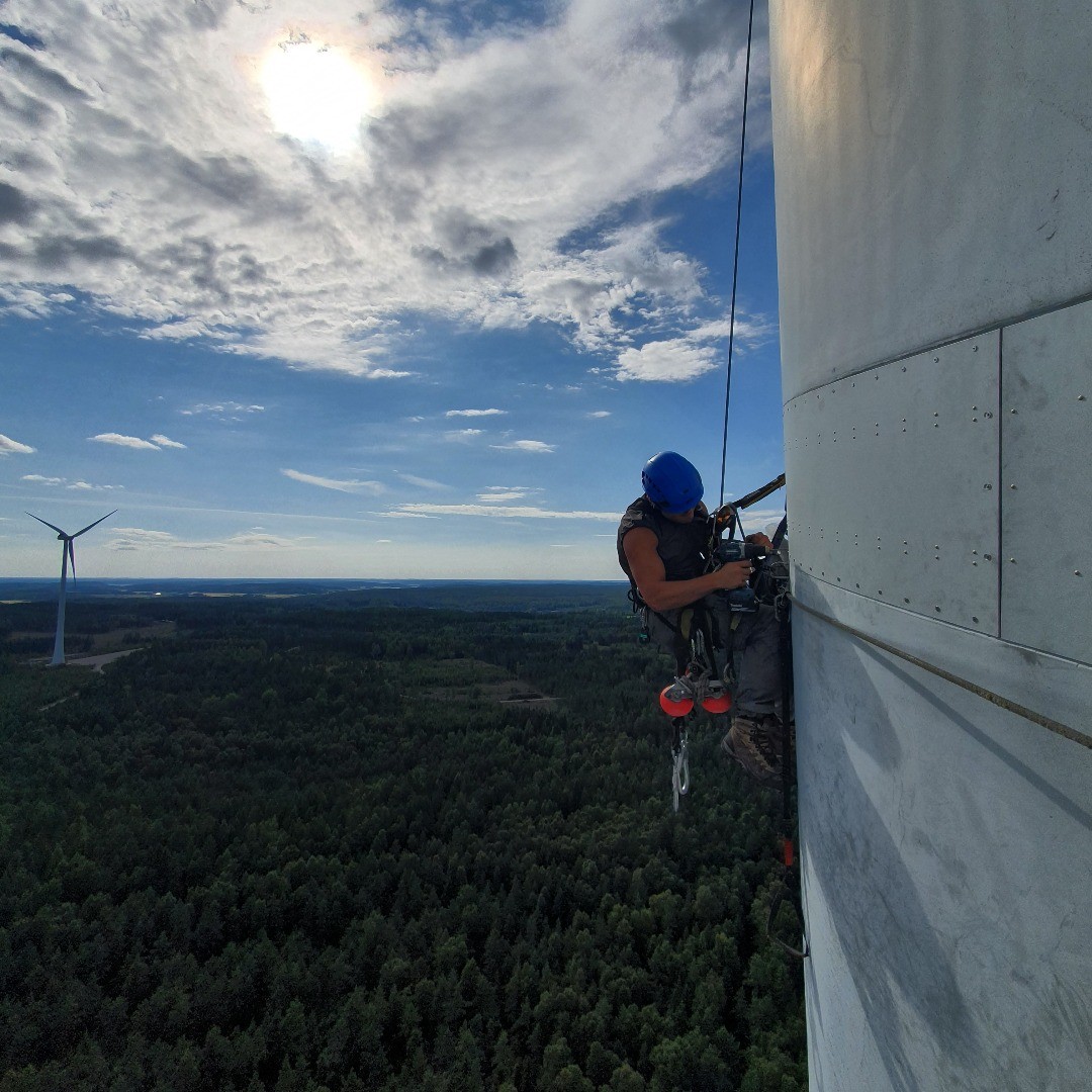 Rope-access technician in helmet and harness working on a wind turbine tower high above a forested landscape.