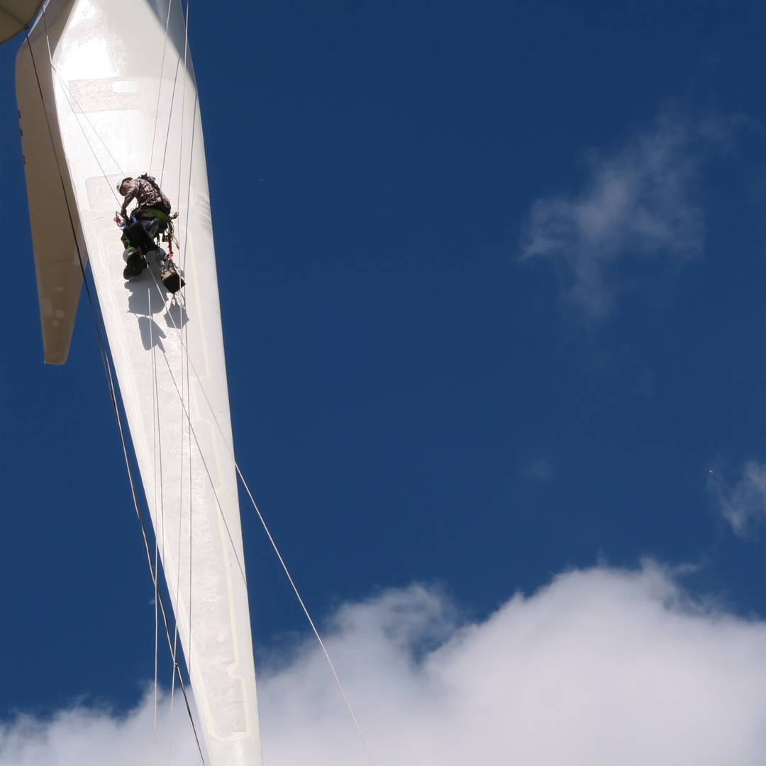 Technician suspended by ropes on a wind turbine blade against a blue sky with clouds, performing maintenance work.