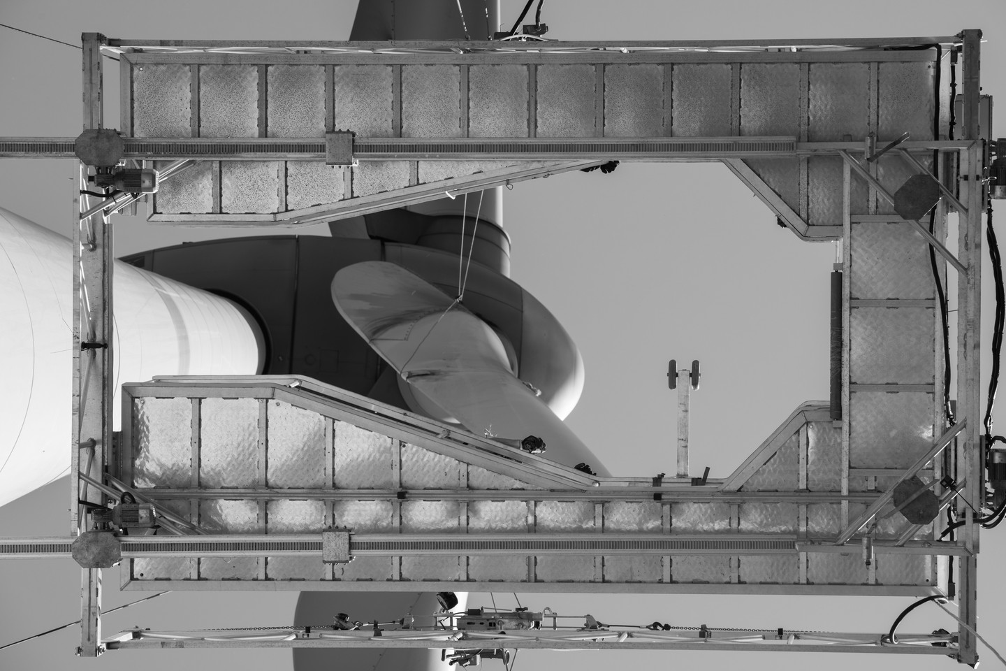 Black and white low-angle view of a wind turbine nacelle and blade seen through a rectangular metal maintenance platform frame.
