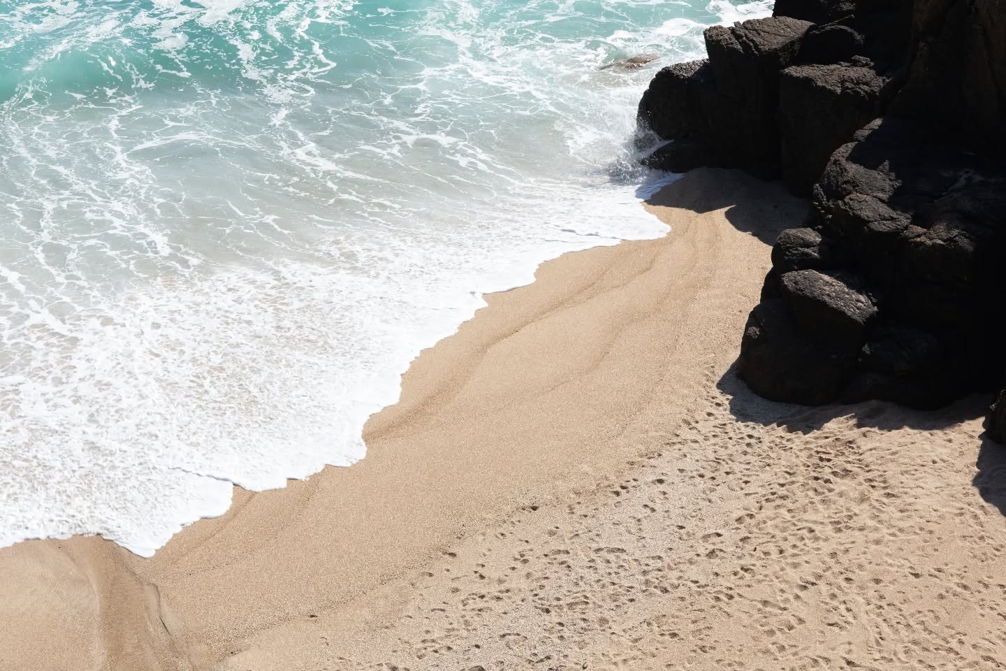 Aerial view of turquoise waves meeting a sandy beach beside dark rocky cliffs, with footprints in the sand.