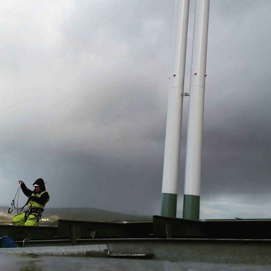 Worker in green safety gear and harness stands on a platform near two tall white poles under a stormy gray sky.