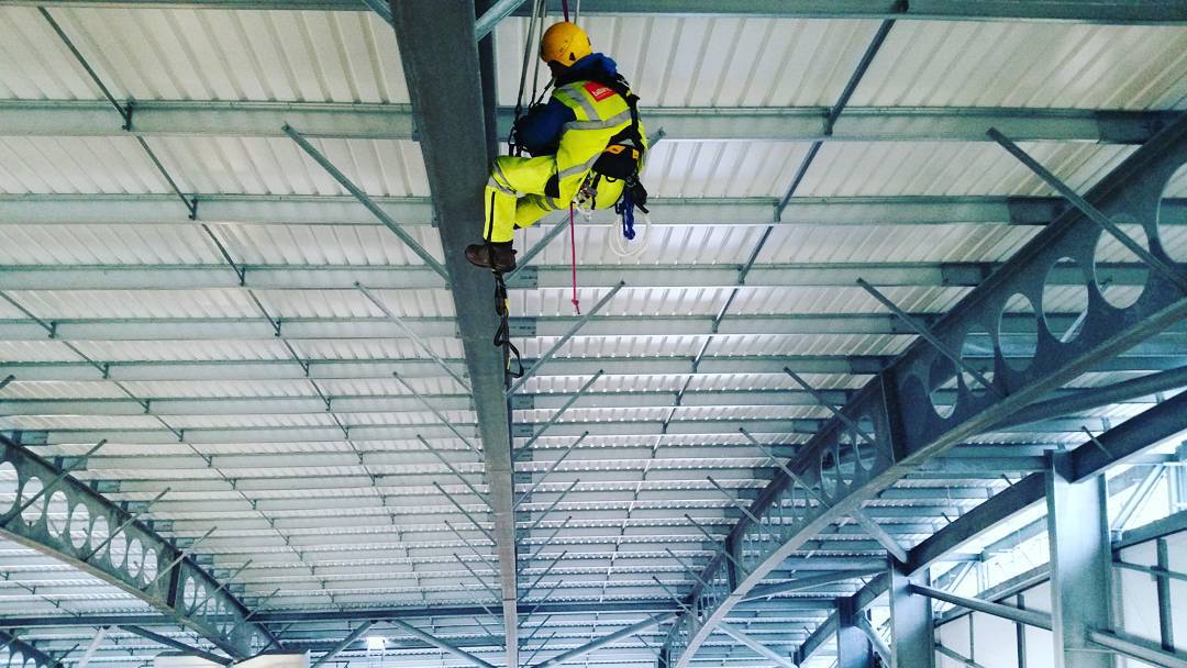 Worker in yellow safety suit and helmet suspended by rope from steel beam under industrial roof structure.